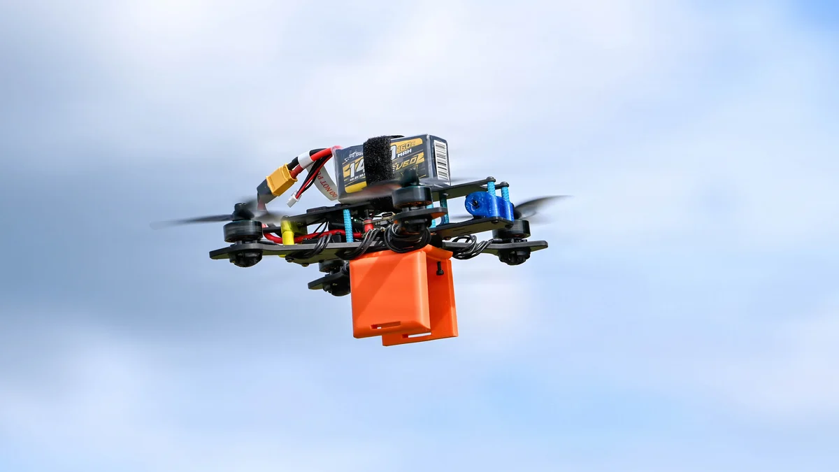 A small quadcopter drone with an orange payload box in flight against a cloudy sky.