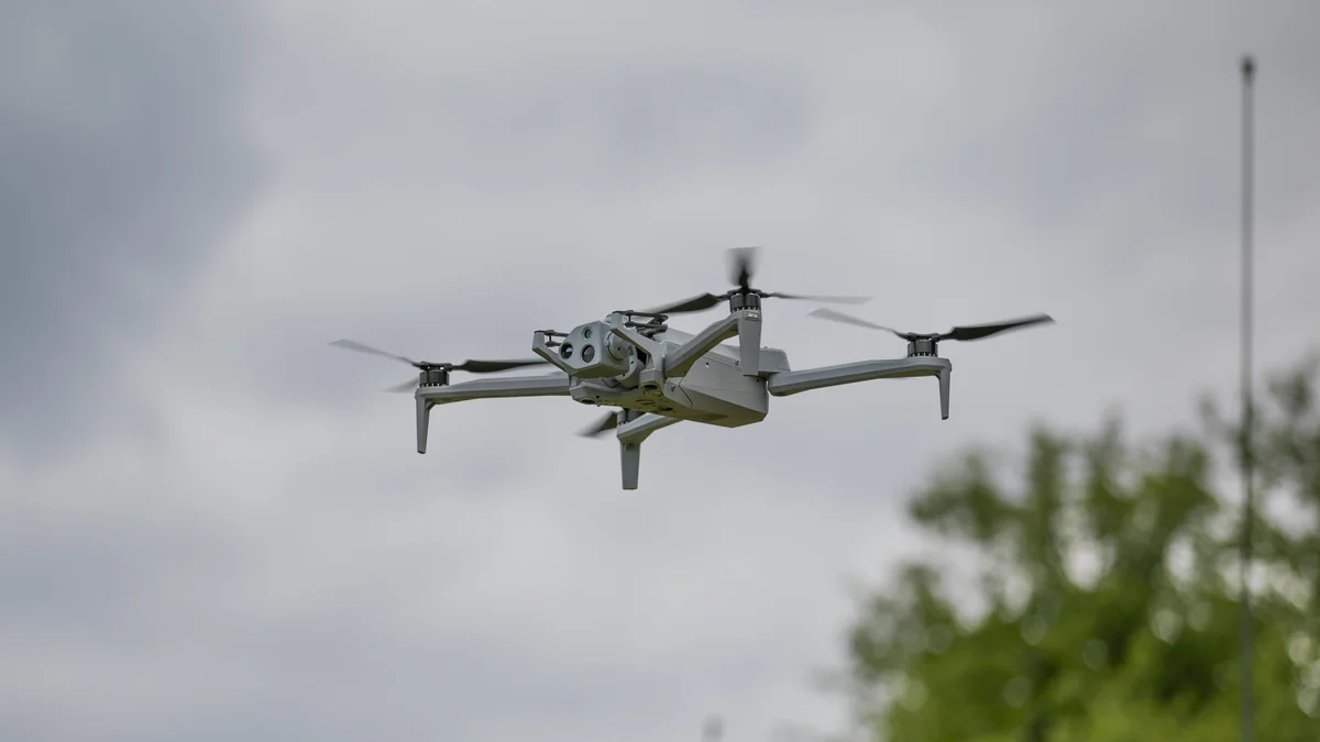 A grey quadcopter drone with a camera gimbal flying under an overcast sky, with trees behind it.