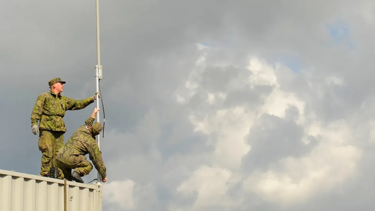 Two soldiers raising a whip antenna mast on the roof of a field shelter under storm clouds.