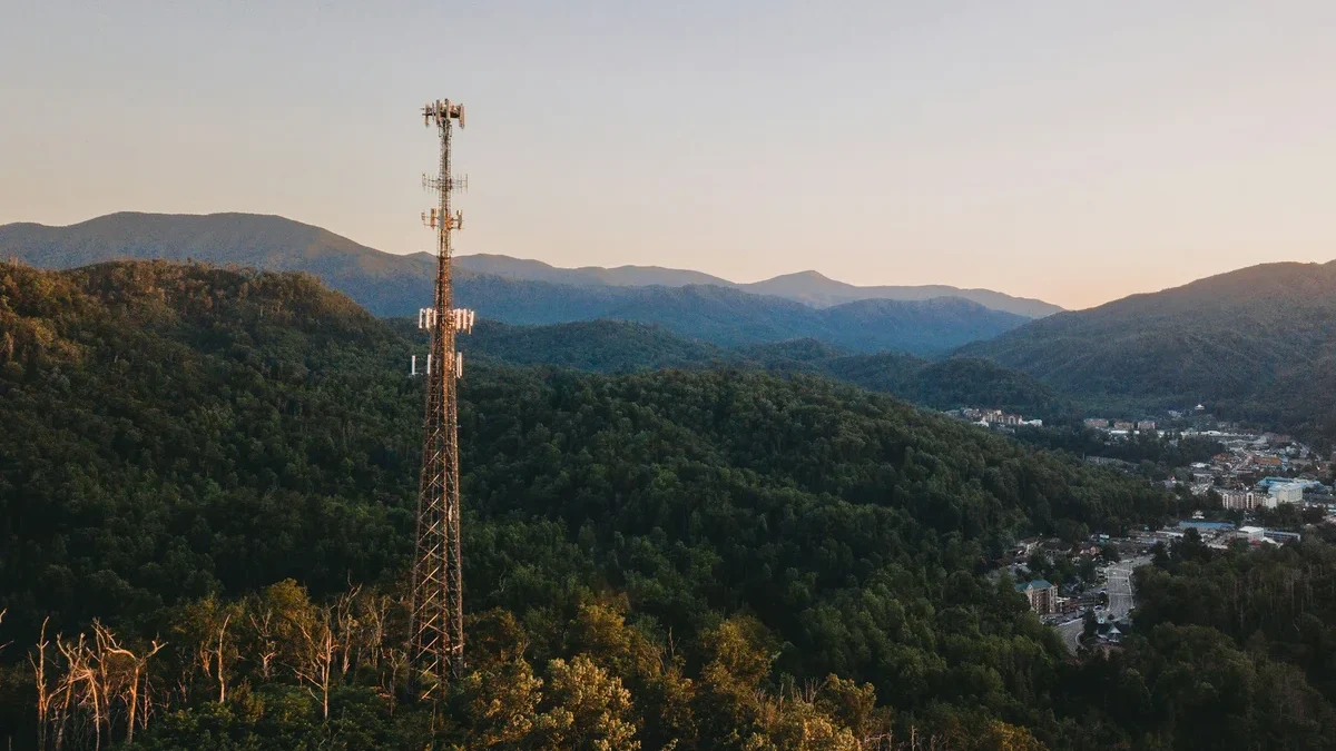 A mobile-network tower on a forested ridge above a valley town at dusk.