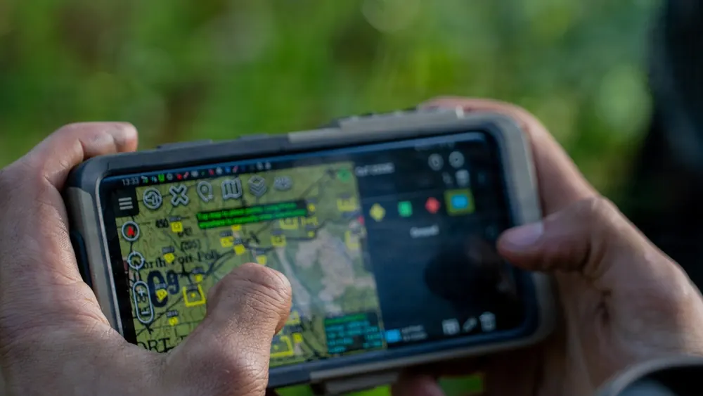 A soldier holding a rugged phone running ATAK, with unit markers on a terrain map.