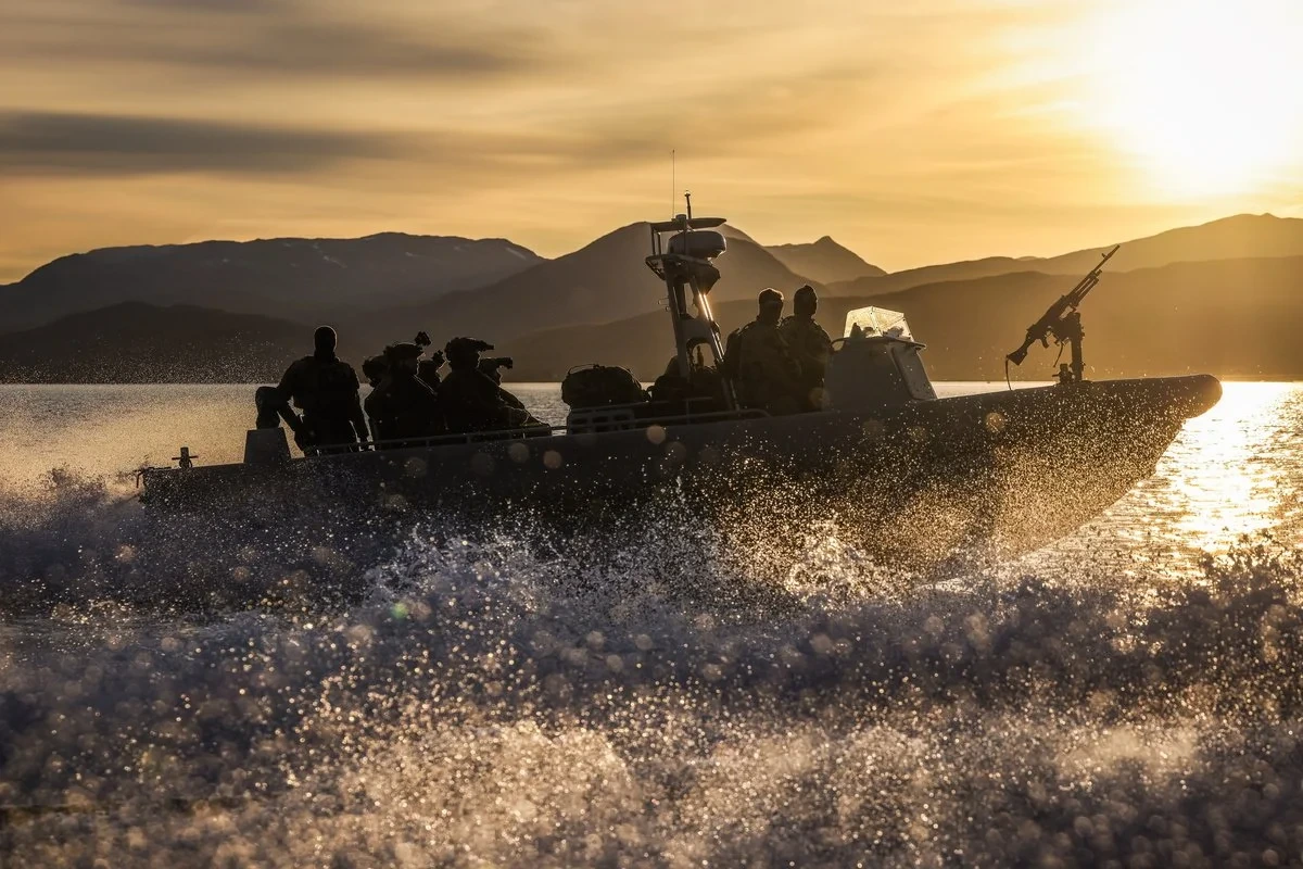 Soldiers in a rigid-hulled boat with a mounted machine gun, speeding across a fjord at sunset.
