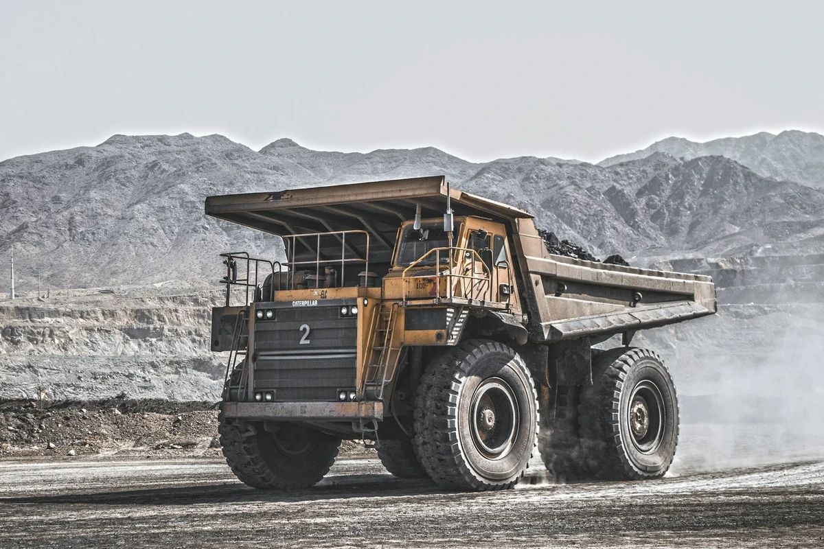 A loaded haul truck driving along a dusty mine road, with the terraced pit walls and mountains behind it.